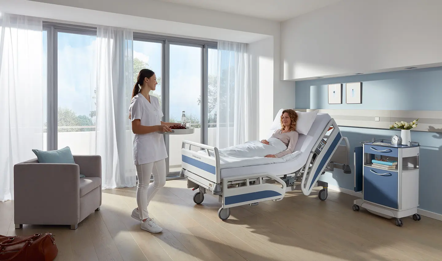 Cama hospitalar Stiegelmeyer Evario com encosto elevado em quarto de internação, paciente em posição semirrecumbente e profissional de enfermagem ao lado do leito