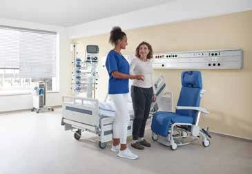 Fase 3 da mobilização precoce: paciente em pé junto ao leito hospitalar Stiegelmeyer conversando com profissional de enfermagem, com poltrona de repouso posicionada ao lado da cama